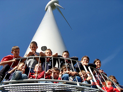 Un groupe d'enfants se tient sur une plate-forme sous une grande éolienne contre un ciel bleu clair, symbolisant la promesse de l'énergie citoyenne en Europe.